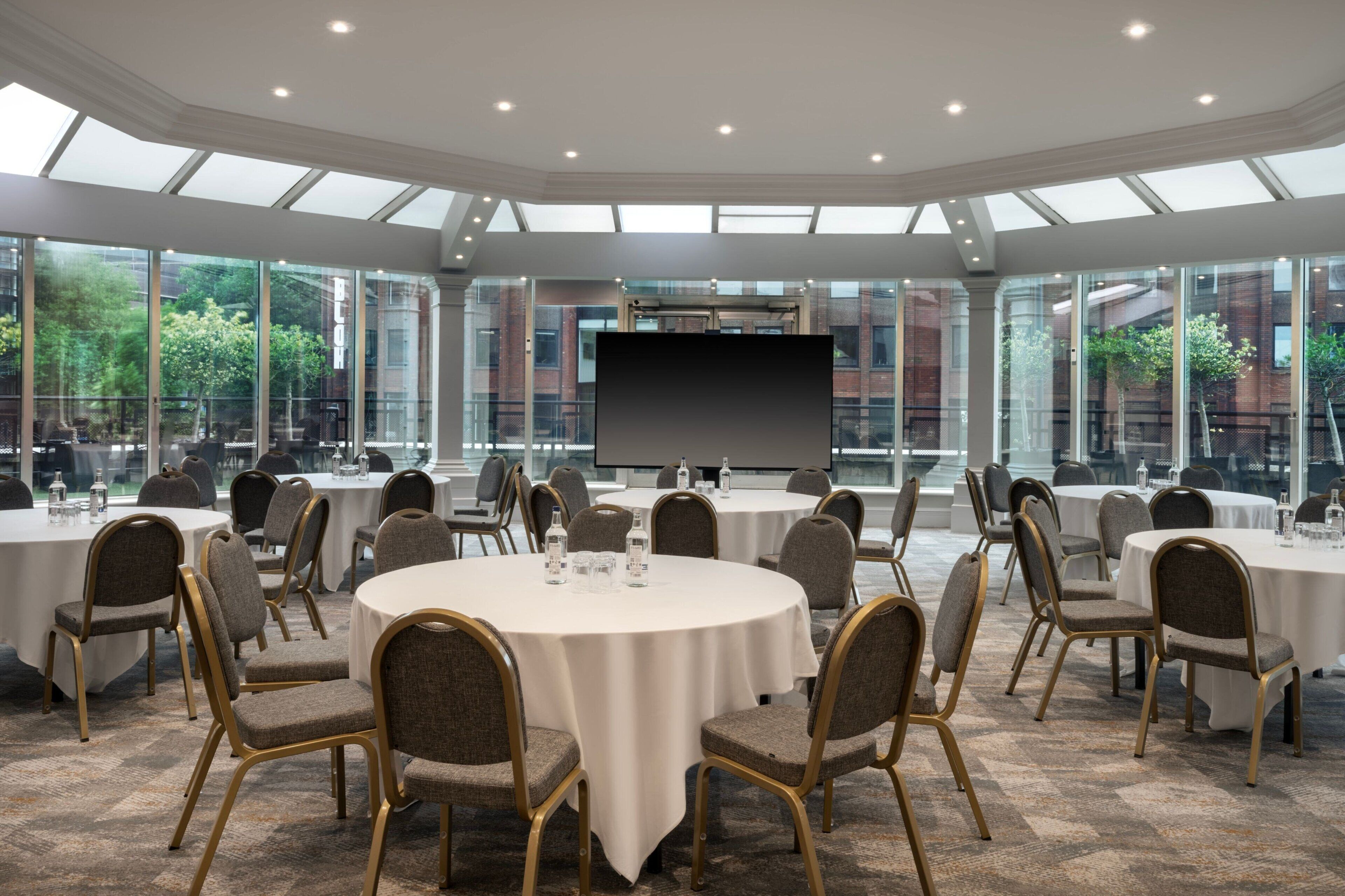 A conference room with round tables and chairs, large windows, and a projection screen.
