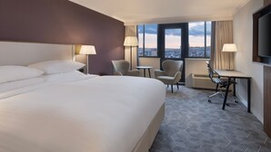 Ein Hotelzimmer mit einem großen Bett, zwei Sesseln, einem Schreibtisch und Blick auf die Stadt durch große Fenster.