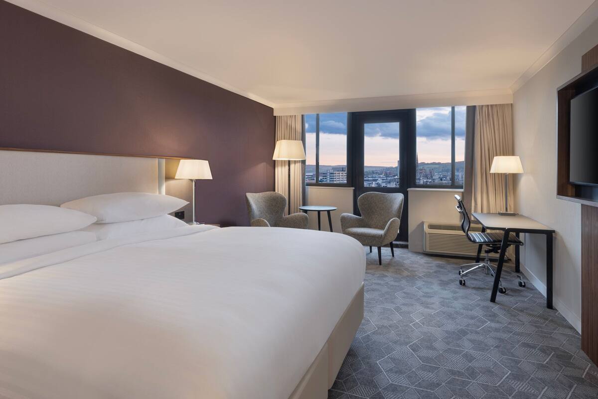 Ein Hotelzimmer mit einem großen Bett, zwei Sesseln, einem Schreibtisch und Blick auf die Stadt durch große Fenster.