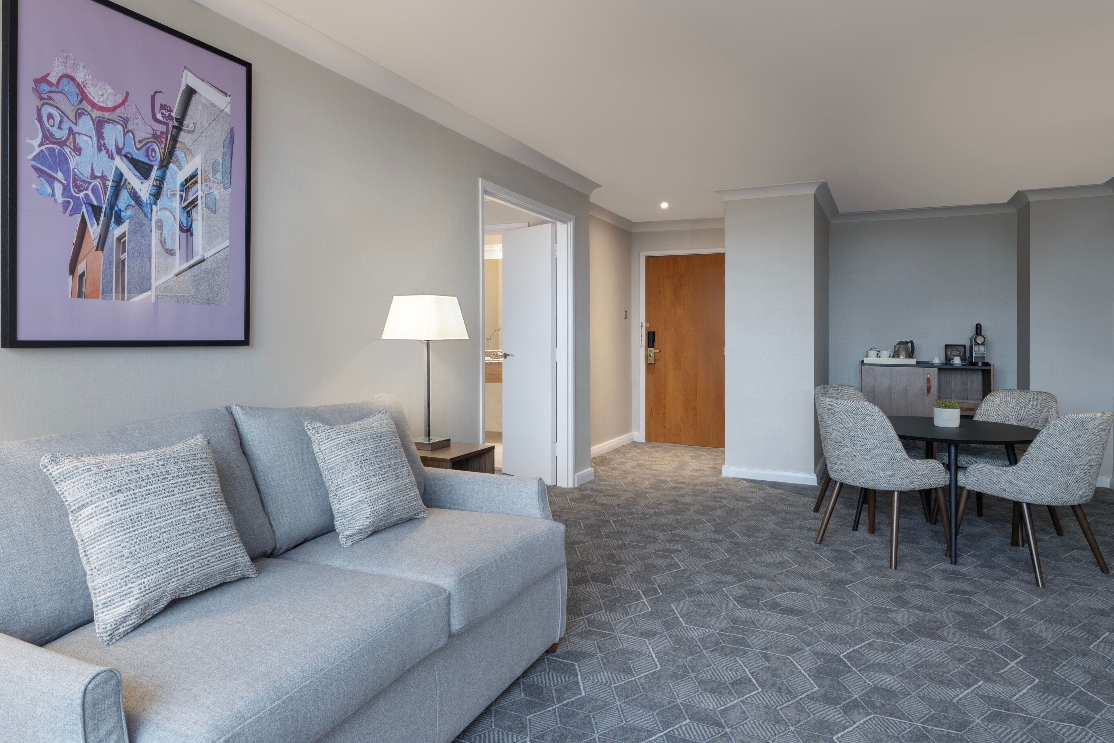 A modern living room with a grey sofa, a dining table with chairs, and a hallway leading to a wooden door.
