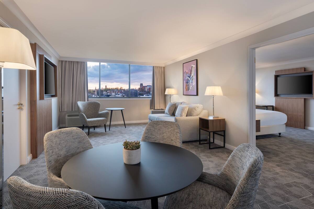 Ein Hotelzimmer mit großem Fenster, einem runden Tisch mit Stühlen, einer Couch, einem Fernseher und einem Bett.