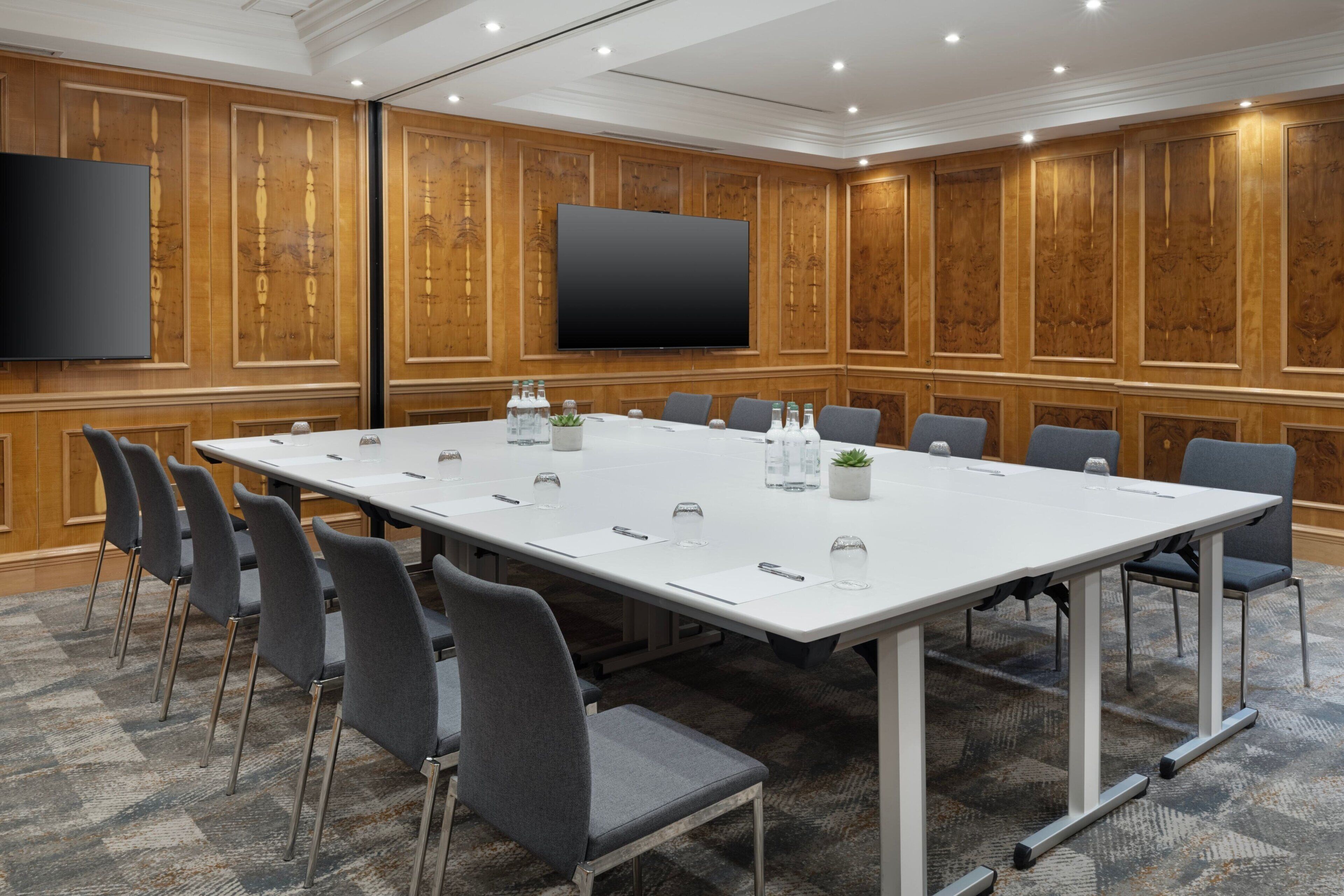 A conference room with a long table, chairs, a flat-screen TV, and wooden paneling.