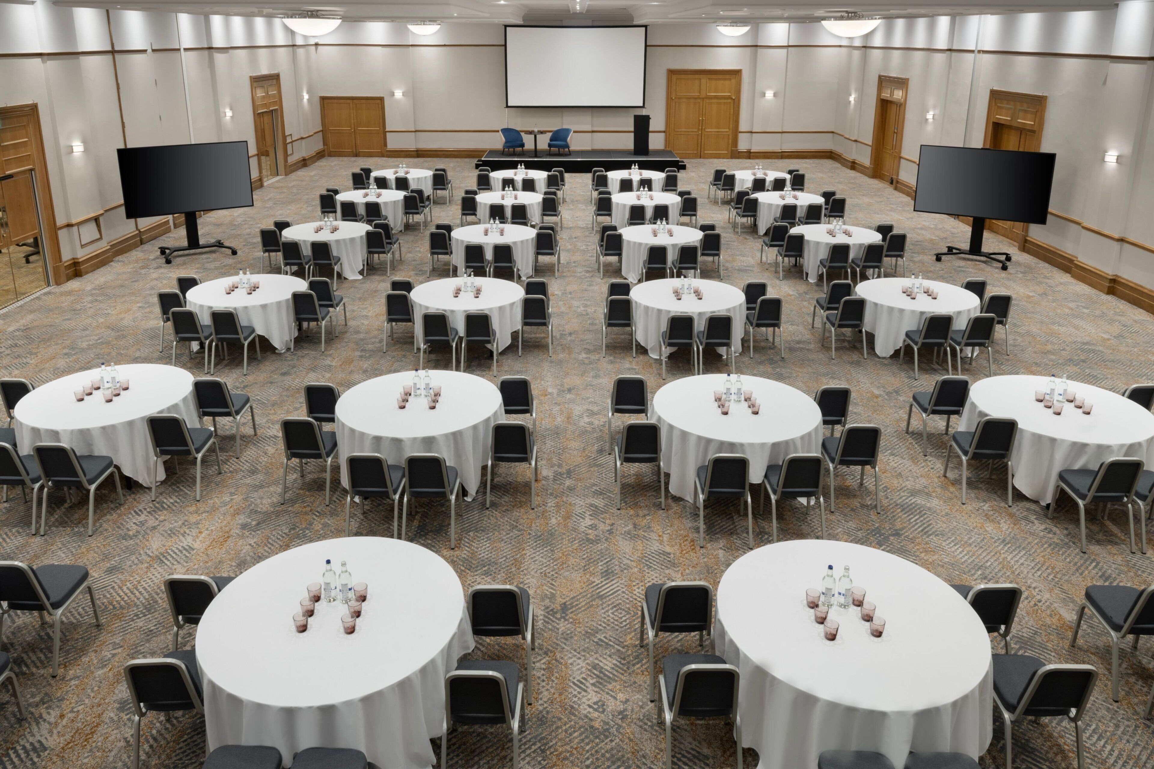 Een congreszaal met meerdere ronde tafels, elk met stoelen en waterflesjes. Er zijn twee grote schermen en een podium aan de voorzijde.