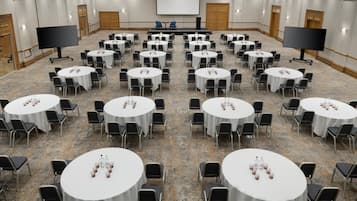 Een congreszaal met meerdere ronde tafels, elk met stoelen en waterflesjes. Er zijn twee grote schermen en een podium aan de voorzijde.