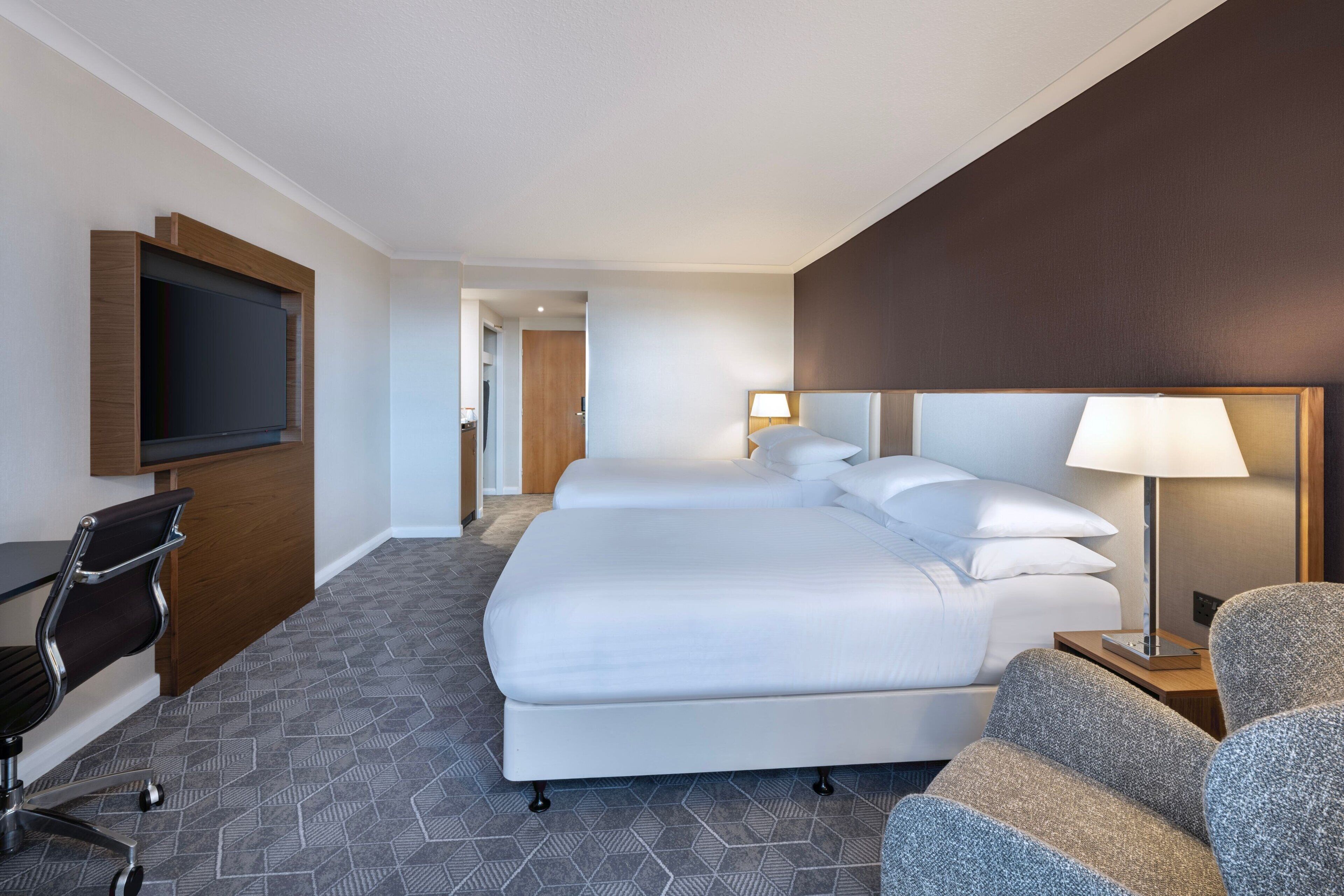 Ein Hotelzimmer mit einem großen Bett, einem Schreibtisch mit Stuhl, einem an der Wand befestigten Fernseher und einer Lampe auf dem Nachttisch.