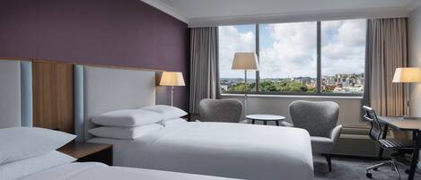 A hotel room with two beds, a desk, a chair, and a view of the city.