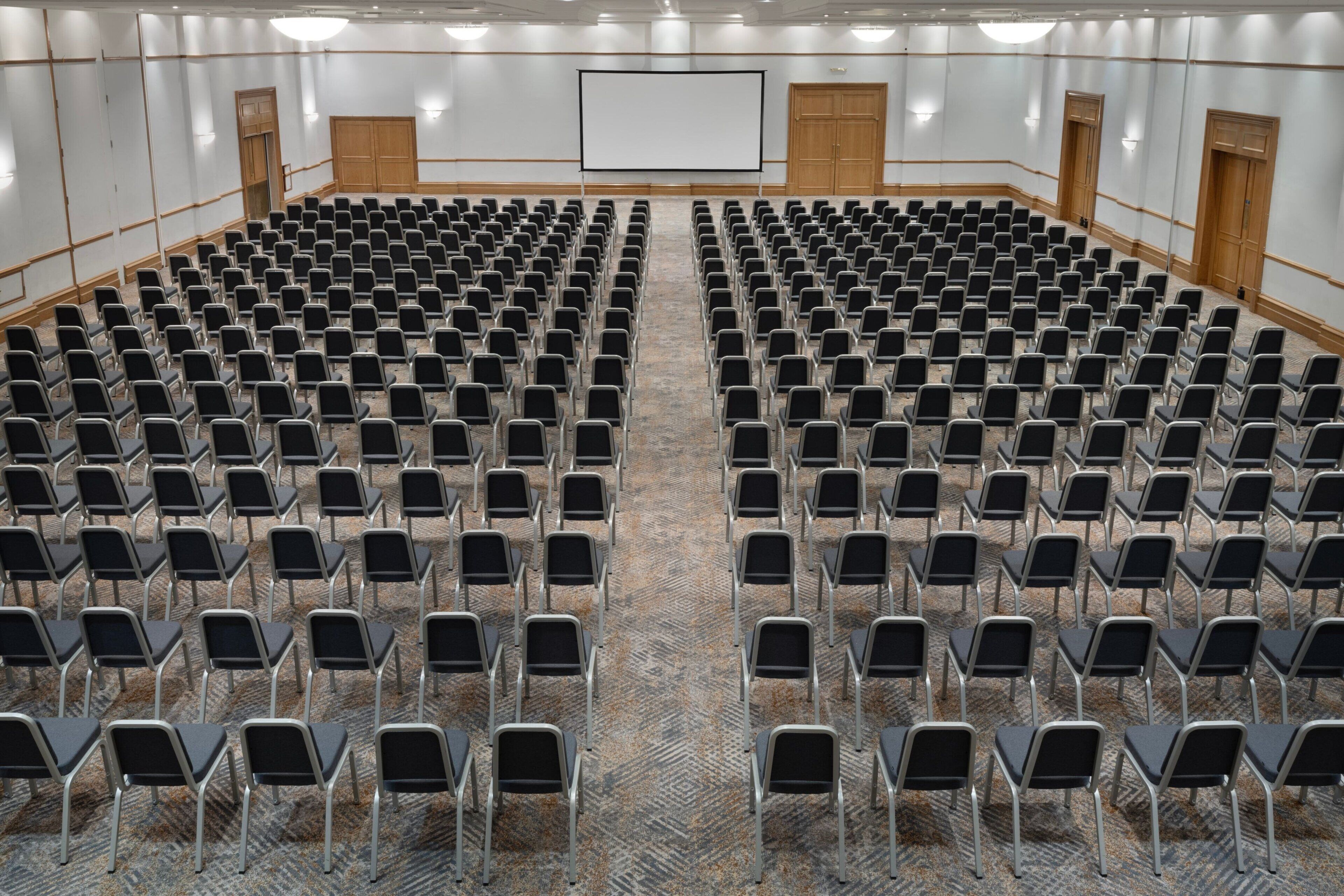 A large conference room with rows of chairs, a projector screen, and wooden doors.