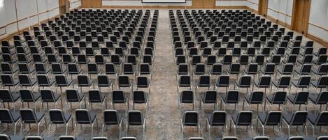 Una grande sala conferenze con file di sedie, uno schermo per proiettore e porte in legno.