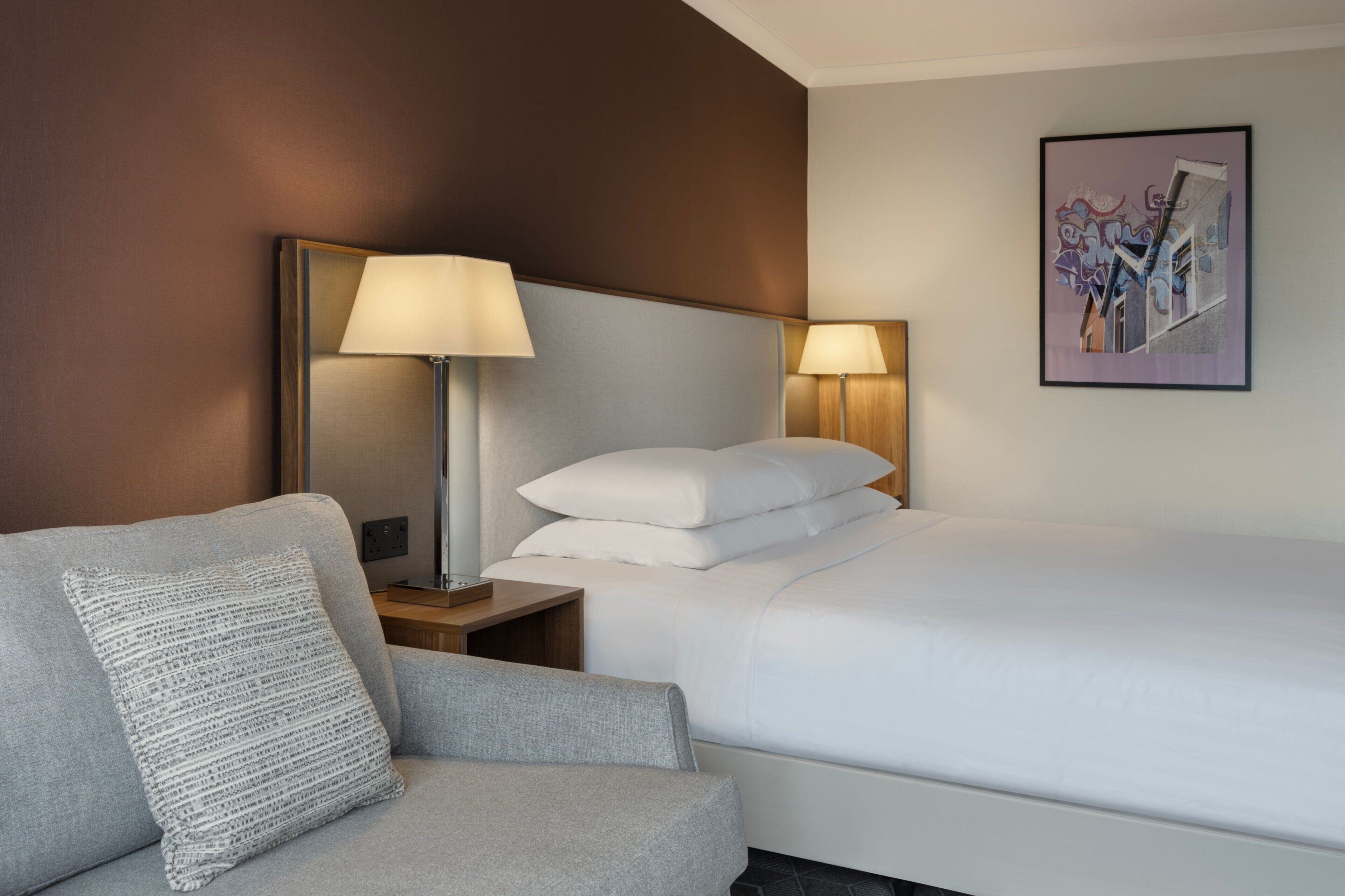 Habitación de hotel con cama, mesita de noche con lámpara, una silla y una obra de arte colgada en la pared.
