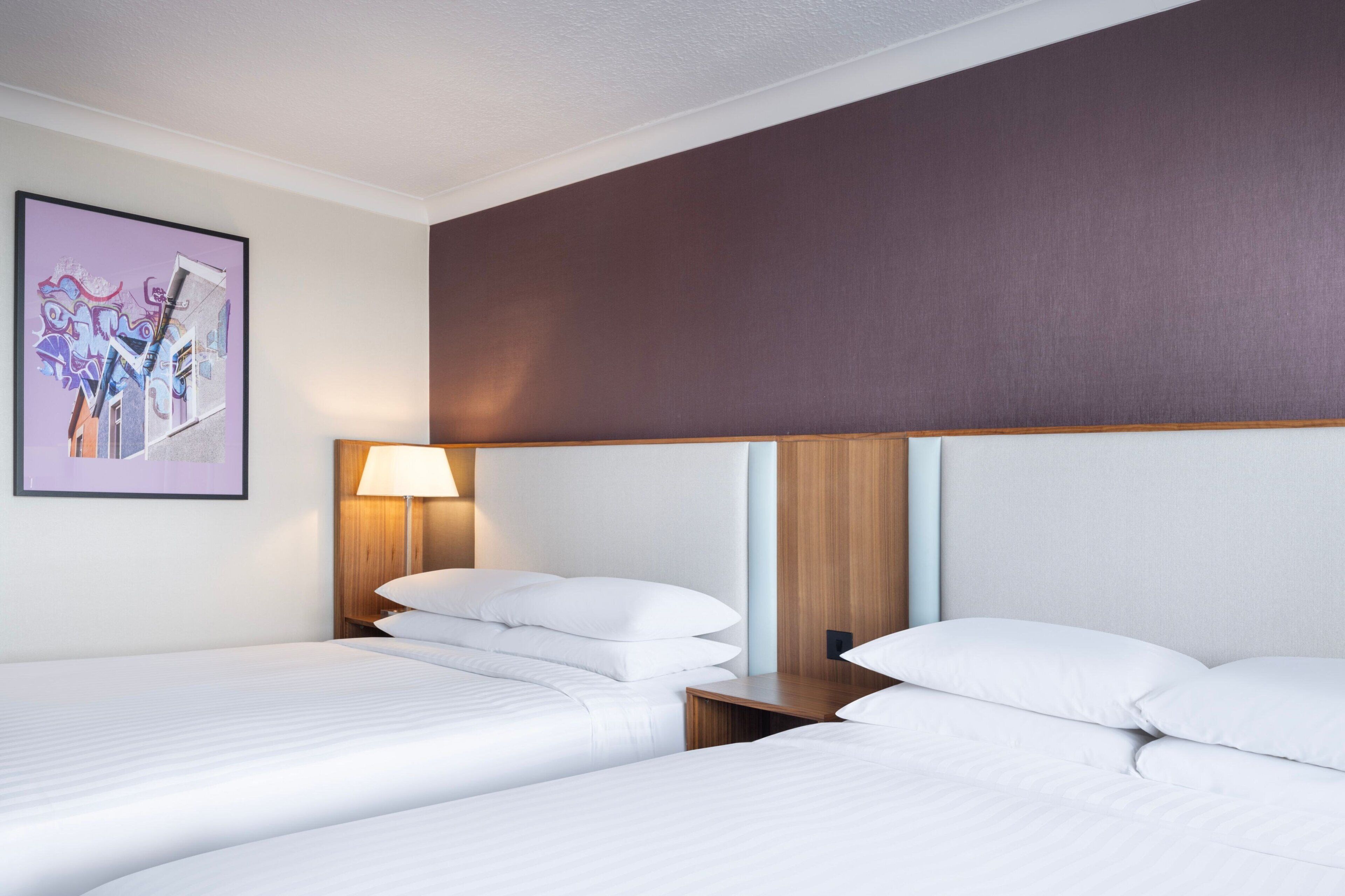 Habitación de hotel con dos camas, cabecero de madera, lámpara de pared y un cuadro enmarcado.