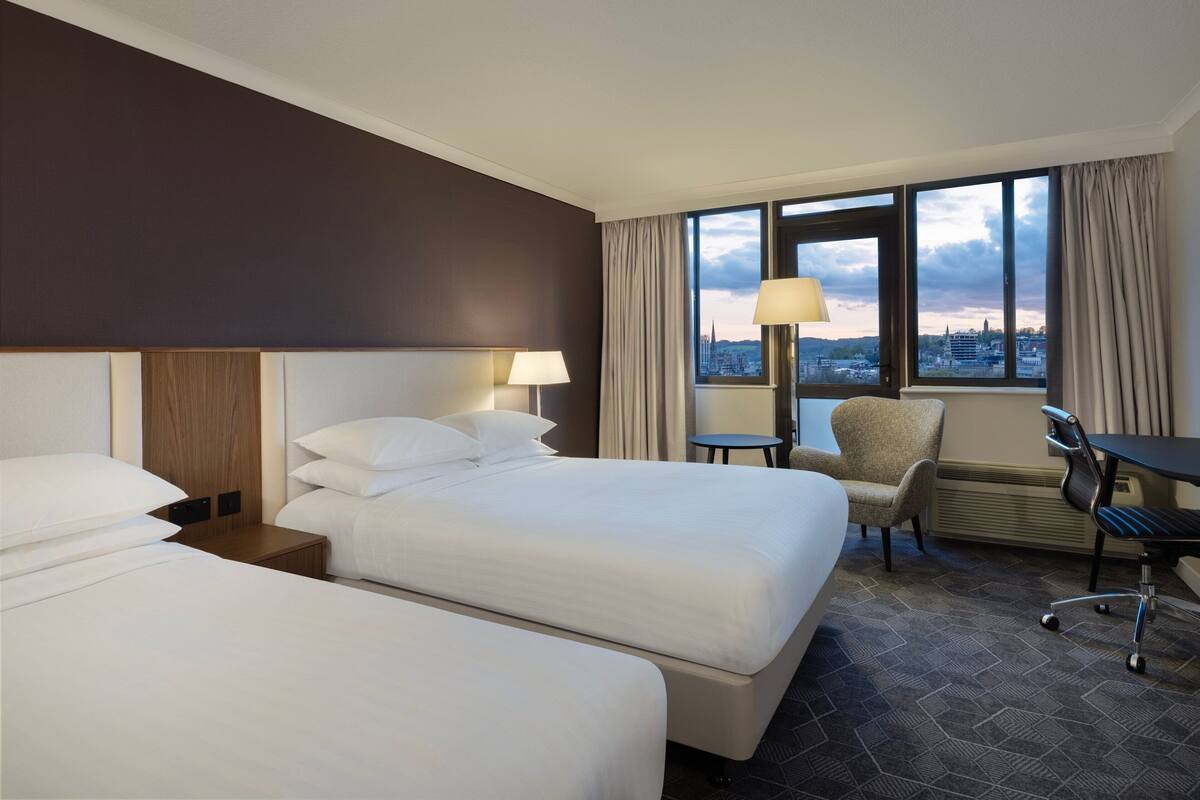 Ein Hotelzimmer mit zwei Betten, einem Schreibtisch, einem Stuhl und Blick auf die Stadt.