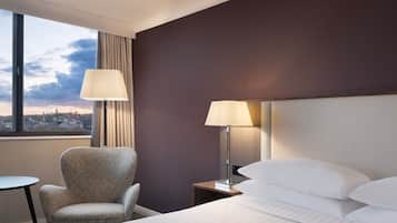 A hotel room with a bed, two bedside lamps, a chair, a small table, and a view of the city through a large window.