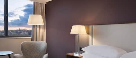 Een hotelkamer met een bed, twee bedlampen, een stoel, een klein tafeltje en uitzicht op de stad door een groot raam.