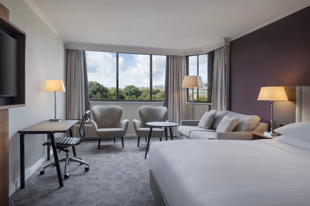 Ein Hotelzimmer mit einem großen Bett, zwei Sesseln, einem kleinen Tisch, einem Schreibtisch mit Stuhl und einem Fernseher.