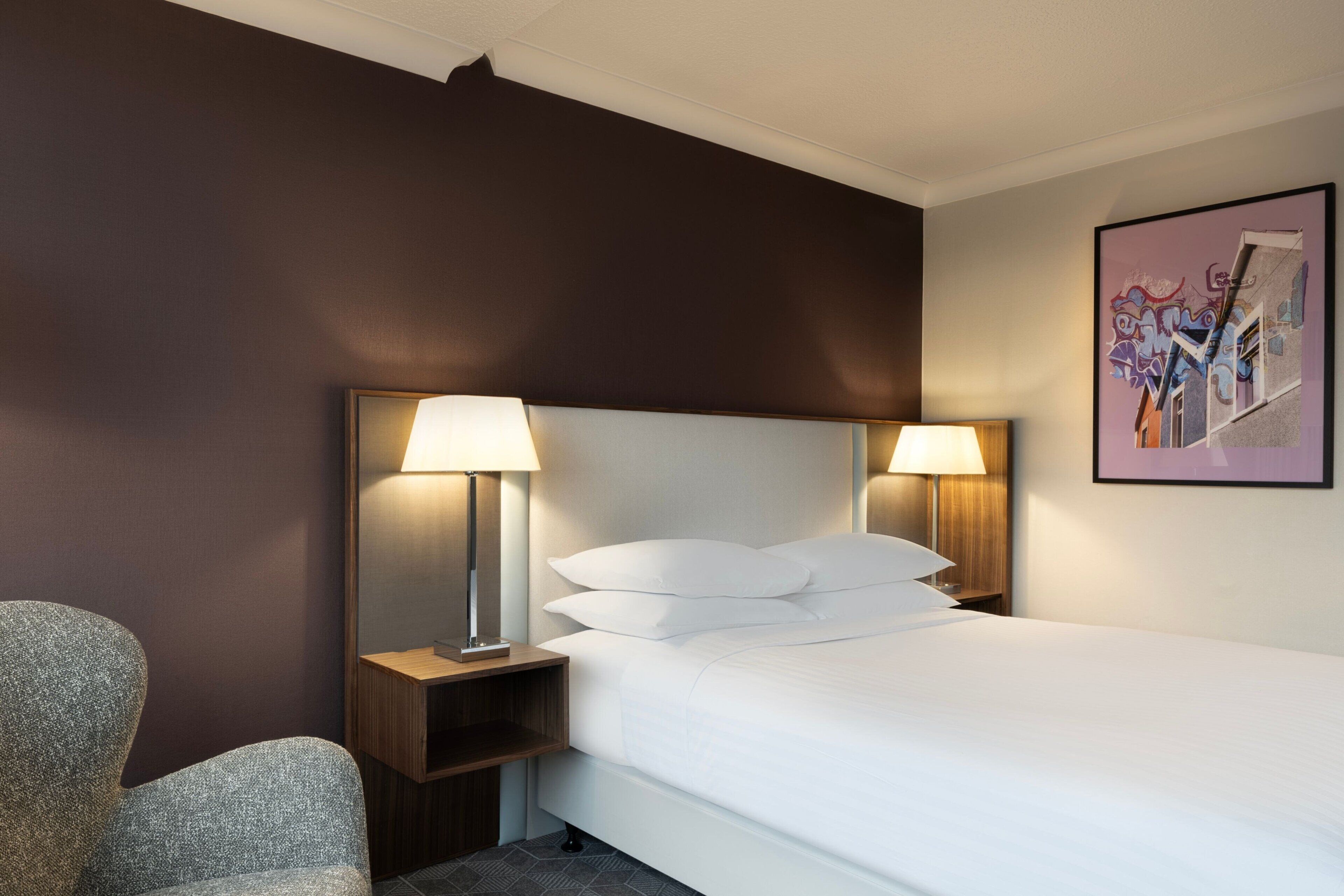 A hotel room with a bed, two bedside lamps, a nightstand, a chair, and a framed artwork on the wall.