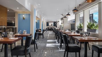A spacious dining area with multiple tables and chairs, large windows, and pendant lighting.
