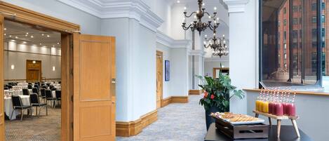A hotel corridor with a carpeted floor, wooden doors, and a chandelier. There is a buffet table with various food items and drinks.