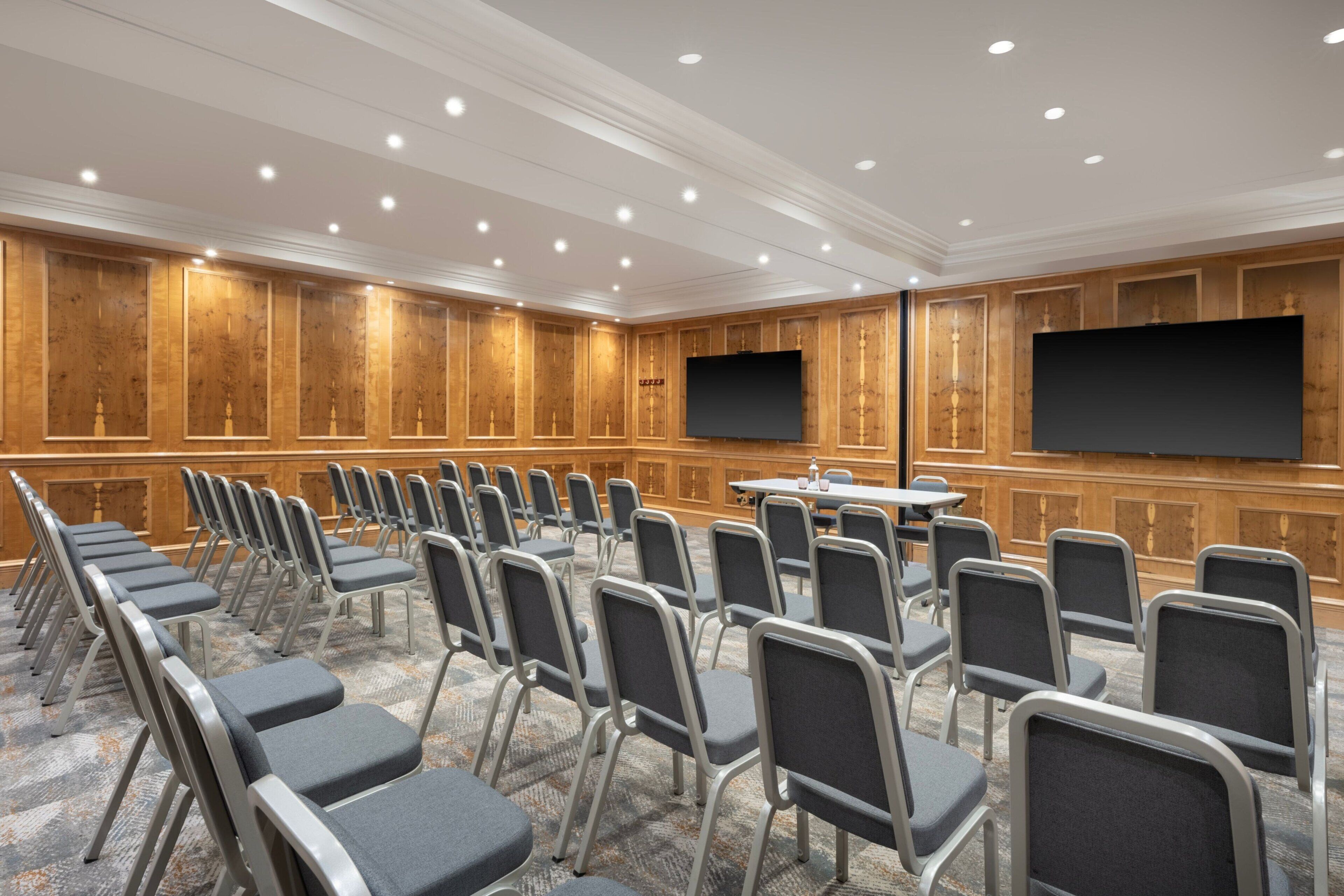 A conference room with rows of chairs, a large table, and two screens on the walls.