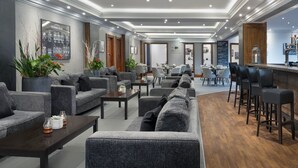 Eine moderne Hotelhalle mit grauen Sofas, einem Couchtisch, einer Bar mit Hockern und einem Essbereich mit Stühlen und Tischen.