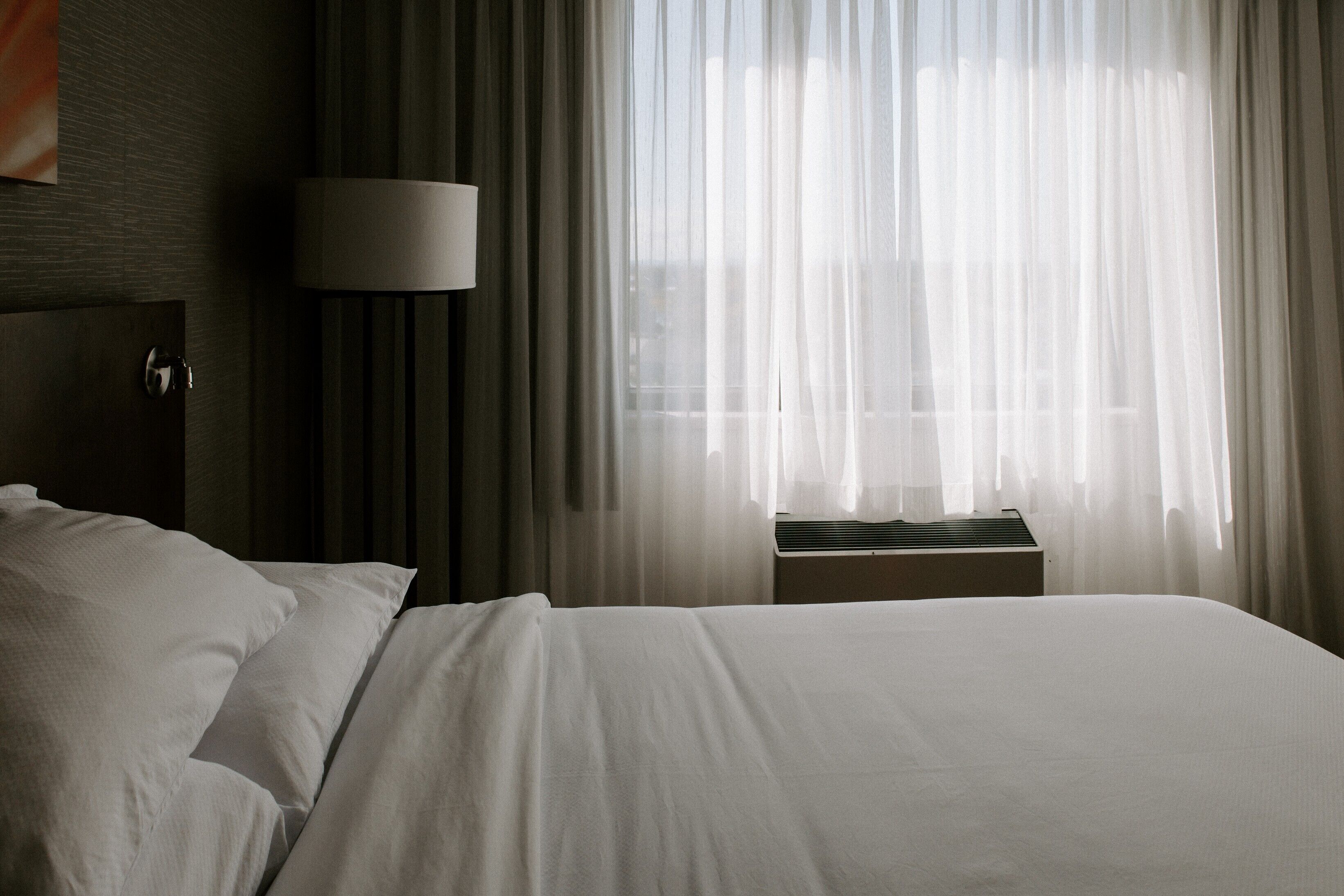 A hotel room with a neatly made bed, a lamp, and sheer curtains.