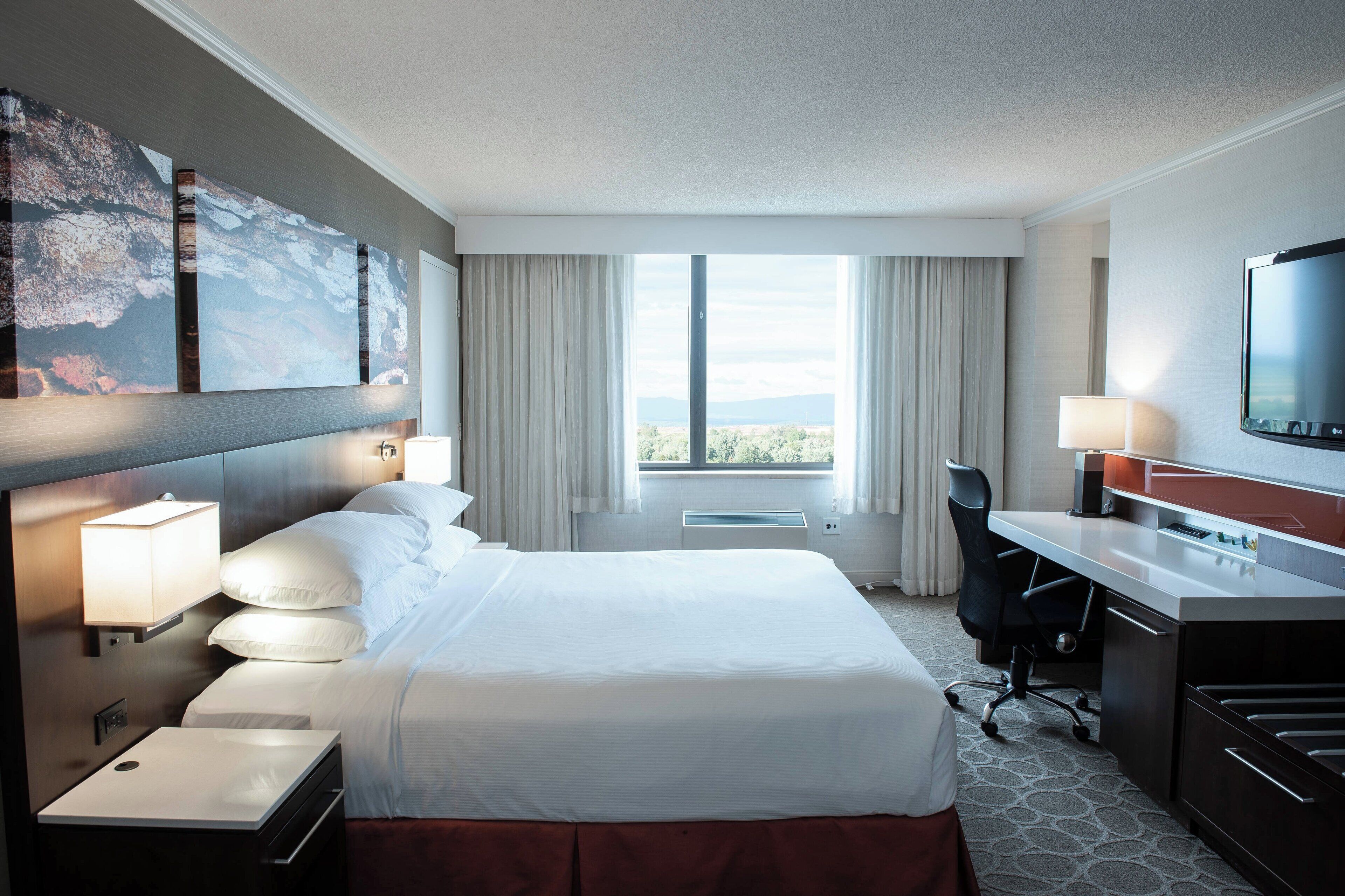 A hotel room with a large bed, a desk with a flat-screen TV, a chair, a desk lamp, a side table, and a large window with curtains.