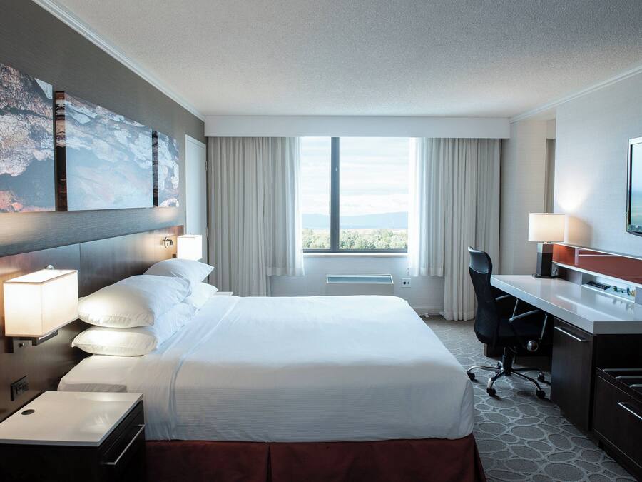 Une chambre d’hôtel avec un grand lit, un bureau doté d’un téléviseur à écran plat, une chaise, une lampe de bureau, une table d’appoint et une grande fenêtre avec des rideaux.