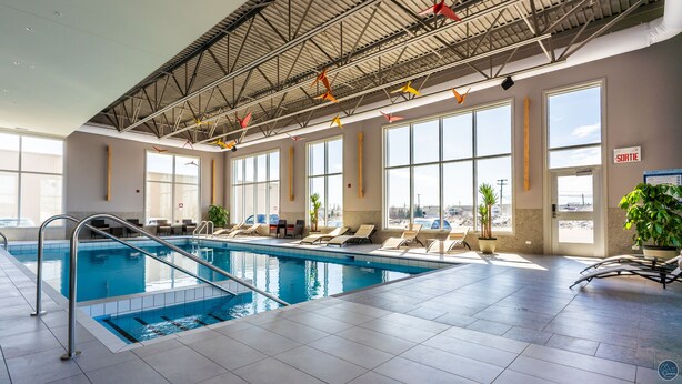 Une piscine intérieure dotée de plusieurs couloirs, de chaises longues et de grandes fenêtres offrant une vue sur l’extérieur.