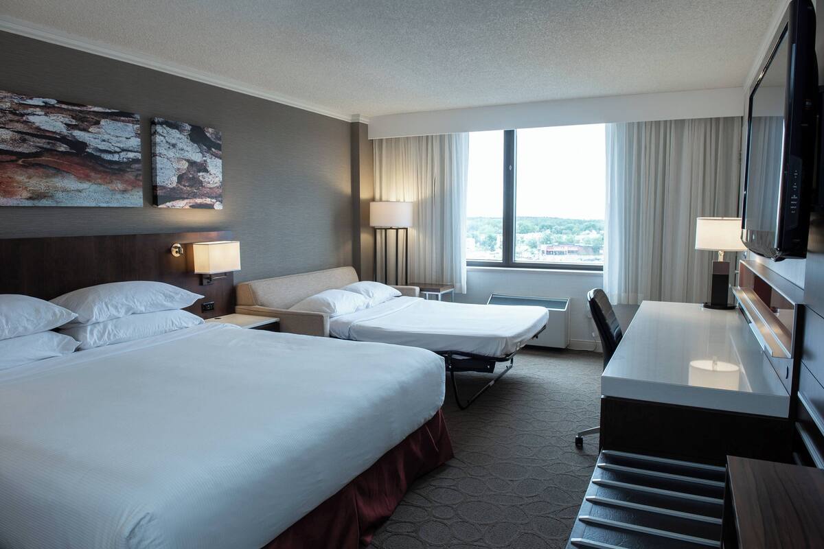 Ein Hotelzimmer mit zwei Betten, einem Schreibtisch, einem Fernseher und einem großen Fenster mit Blick auf die Stadt.