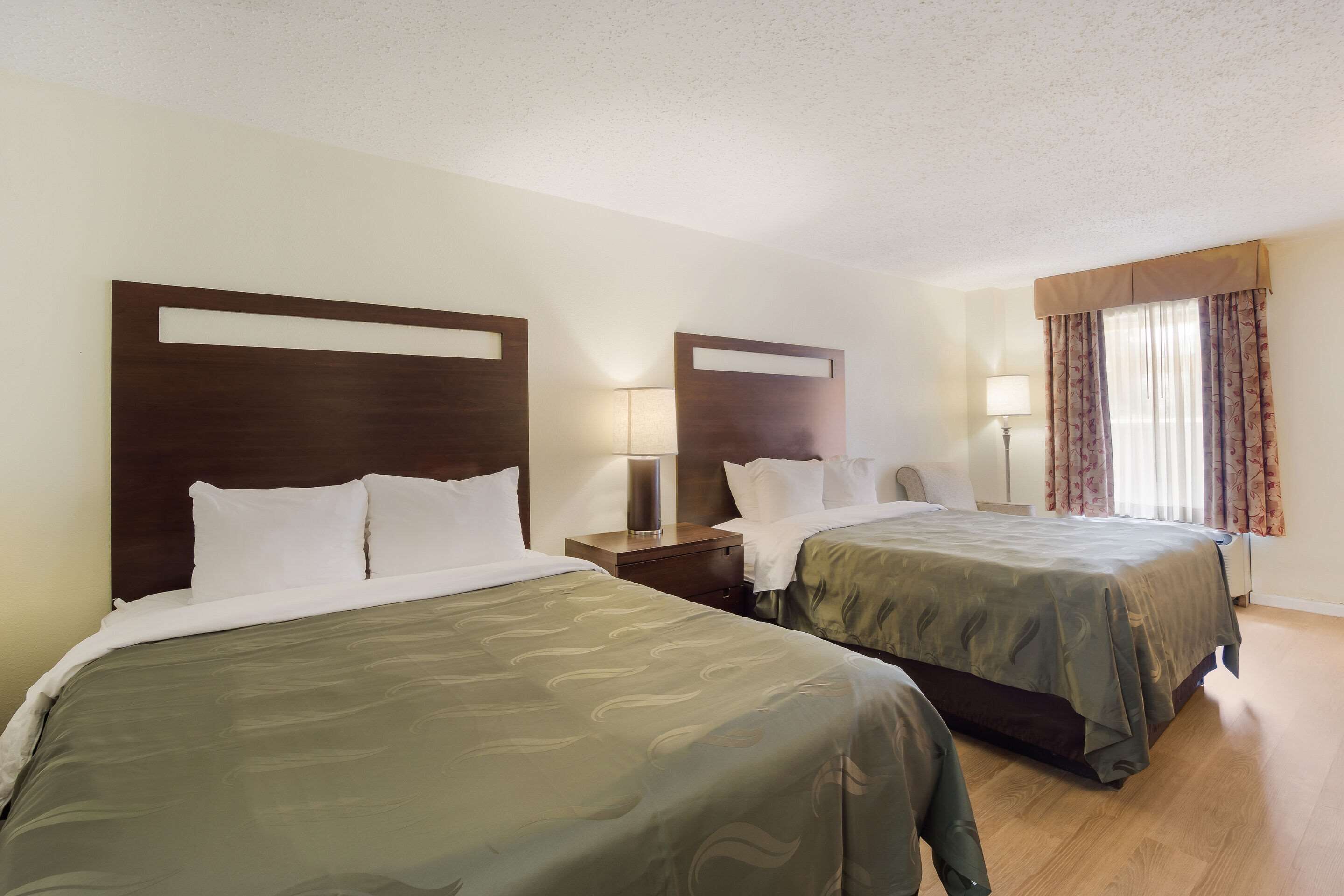 A hotel room with two beds, wooden headboards, bedside lamps, and a window with curtains.