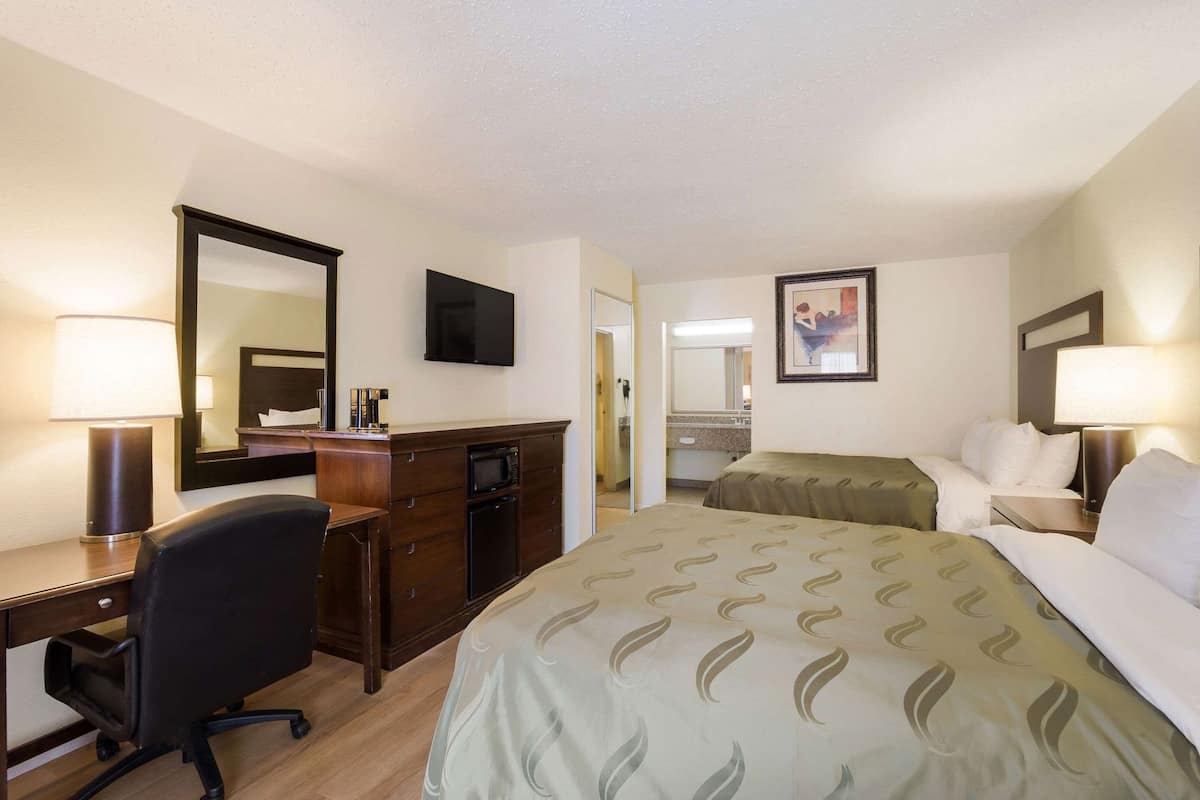 Habitación de hotel con escritorio, dos camas, cómoda, televisor y baño visible a través de una puerta abierta.
