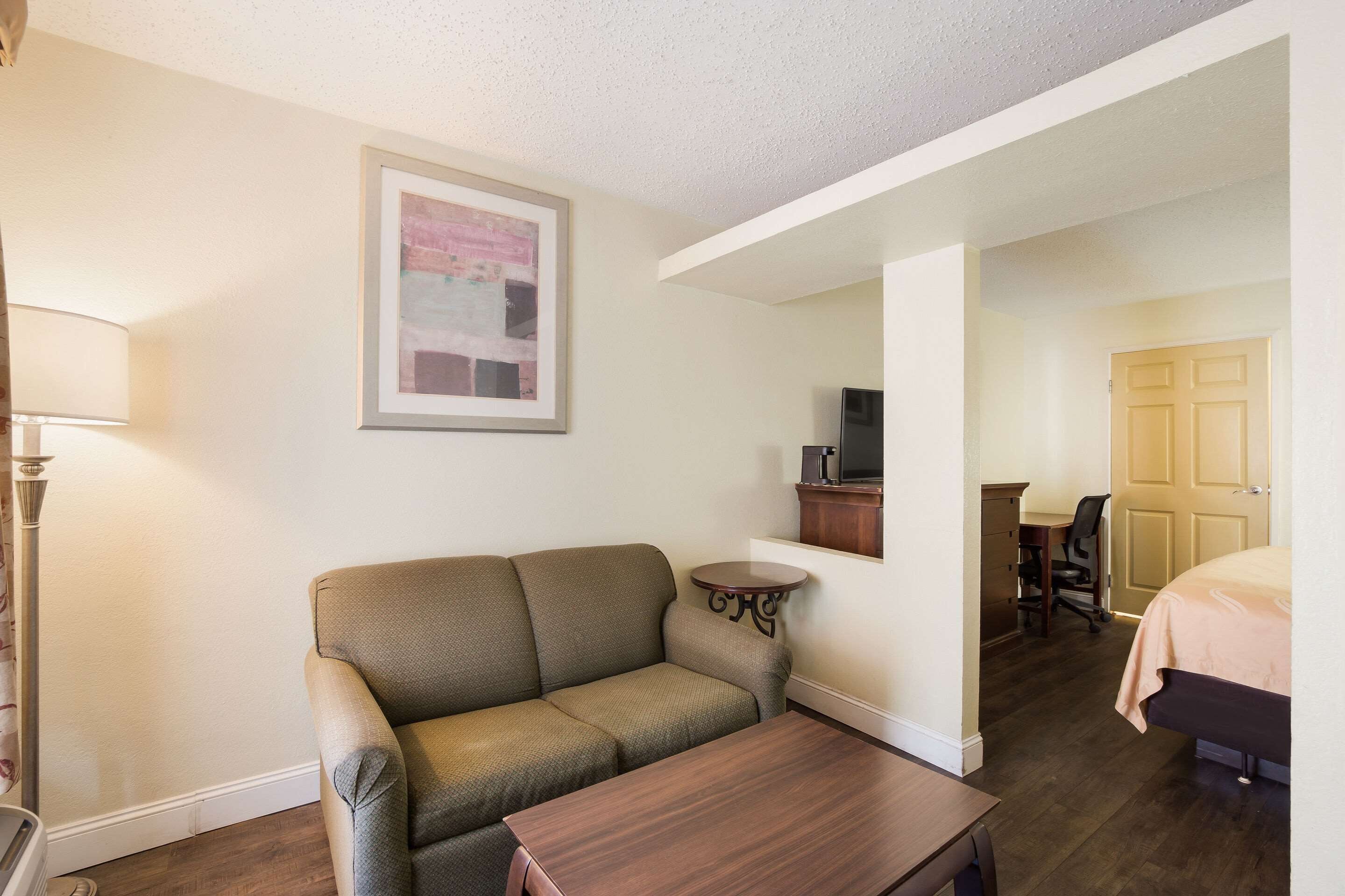 A room with a sofa, a small table, a lamp, a TV, a desk with a chair, and a bed.