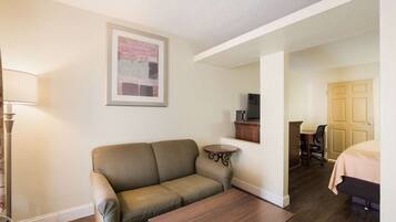 A room with a sofa, a small table, a lamp, a TV, a desk with a chair, and a bed.