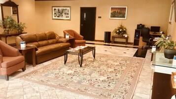 A spacious lobby with a brown leather sofa, a coffee table, and a floral rug. There are two armchairs, a small table with a vase, and a door with a sign.