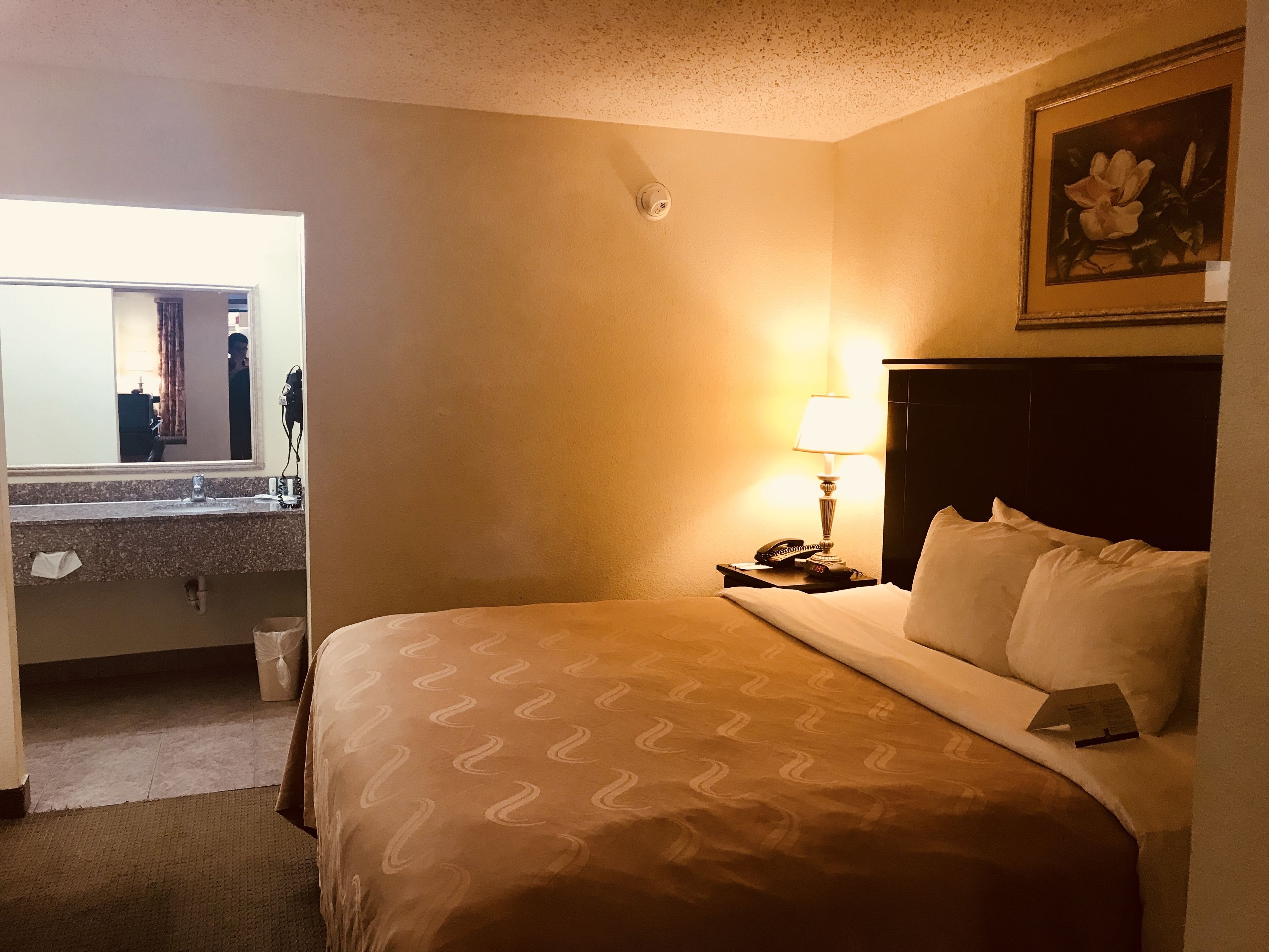 Quarto de hotel com cama, mesa de cabeceira com abajur, um quadro na parede e banheiro visível através de uma porta aberta.
