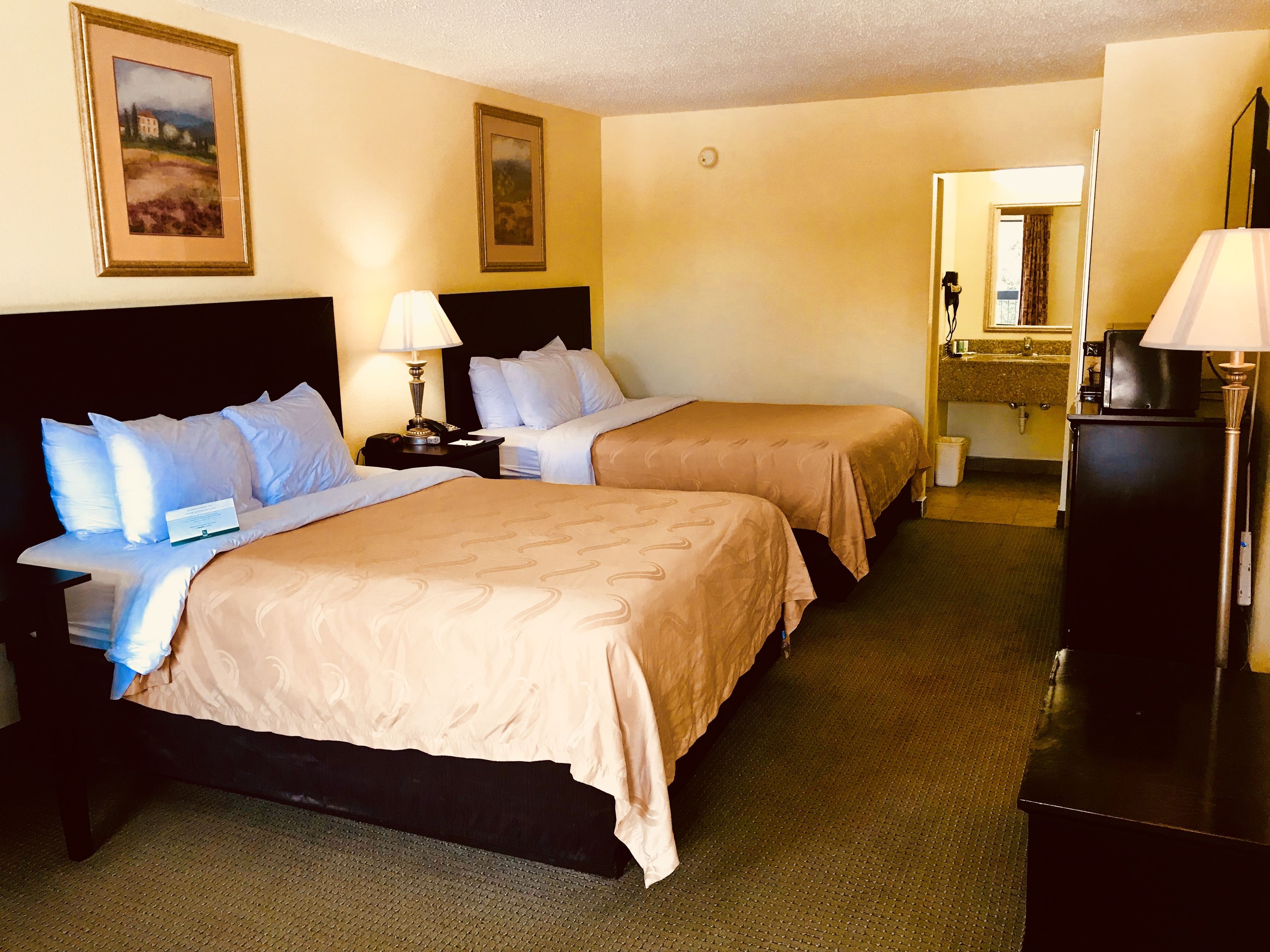 A hotel room with two beds, a nightstand, a lamp, a refrigerator, and a bathroom visible in the background.
