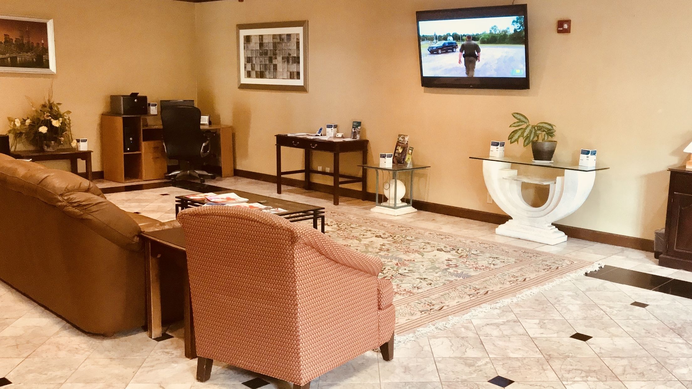 A lobby with a brown sofa, a patterned armchair, a TV, a desk, and a decorative glass table.