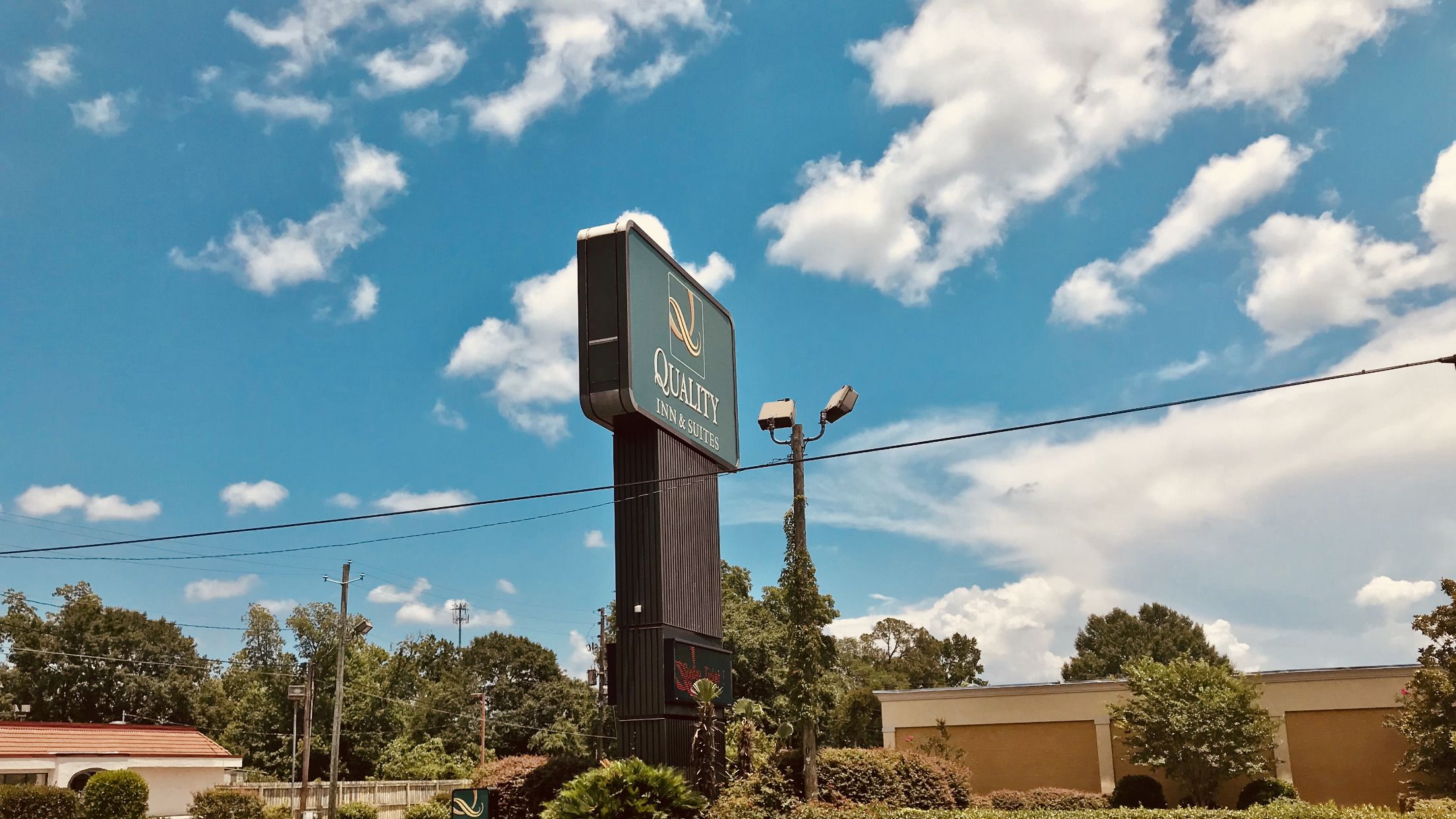 Placa de um hotel chamado "Quality Inn & Suites", com um céu azul claro e nuvens ao fundo.