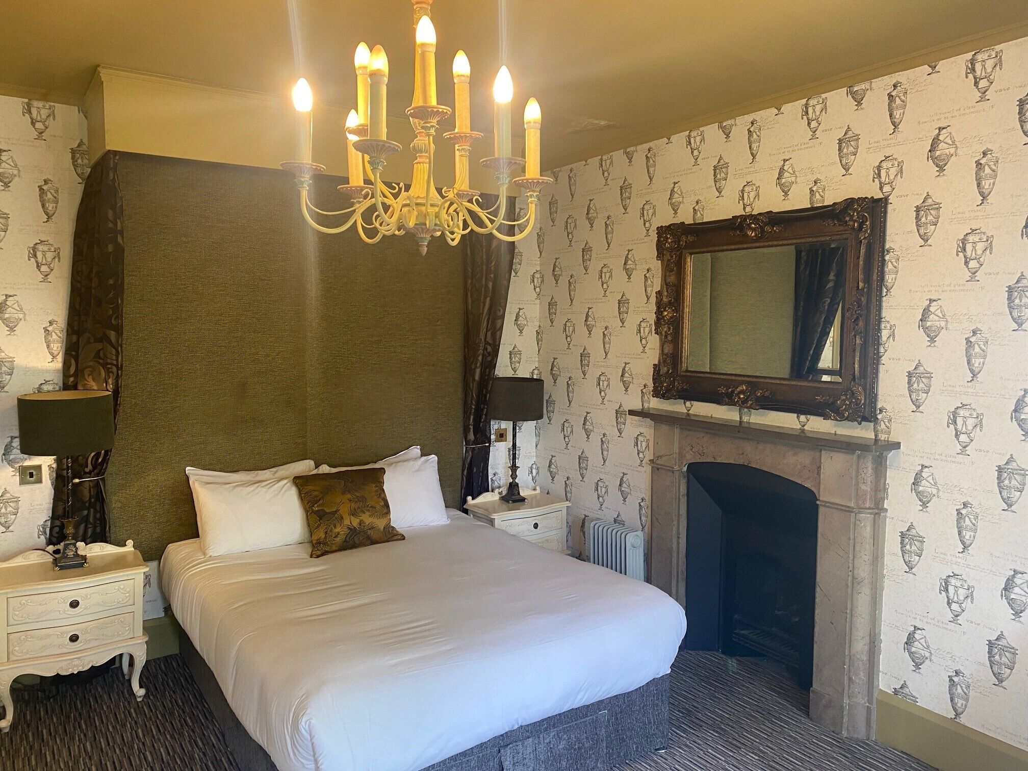 A hotel room with a large bed, a chandelier, a fireplace, and a mirror above it.