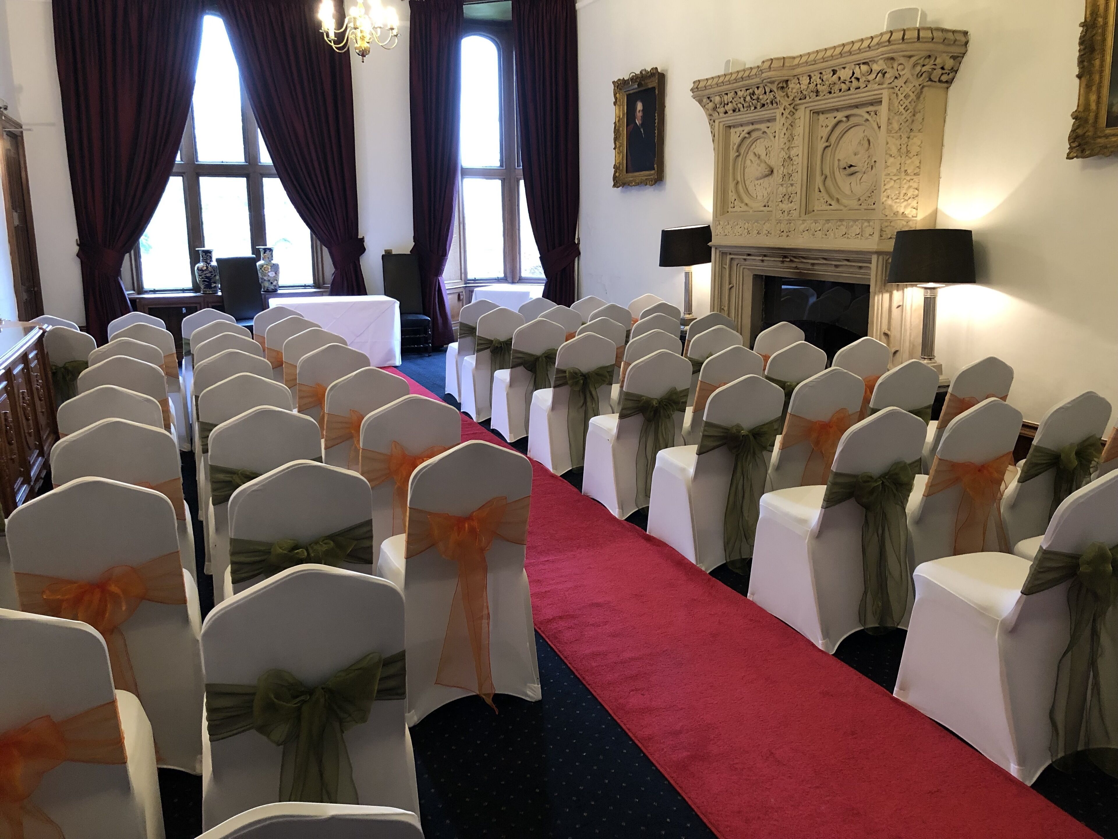 Une salle ornée de rangées de chaises blanches avec des rubans orange, un tapis rouge et une cheminée aux sculptures complexes.