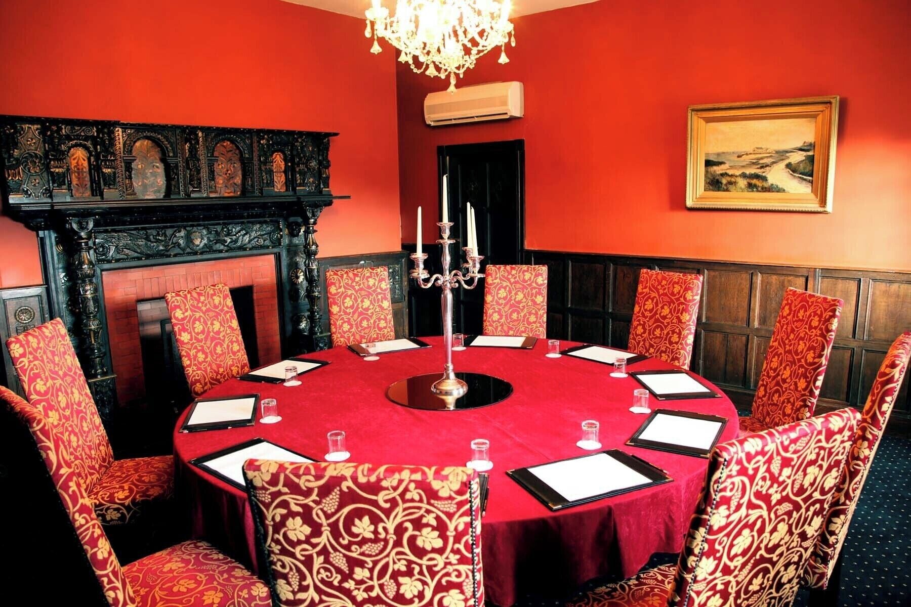 Una mesa redonda con mantel rojo y sillas a juego, una araña y una chimenea.