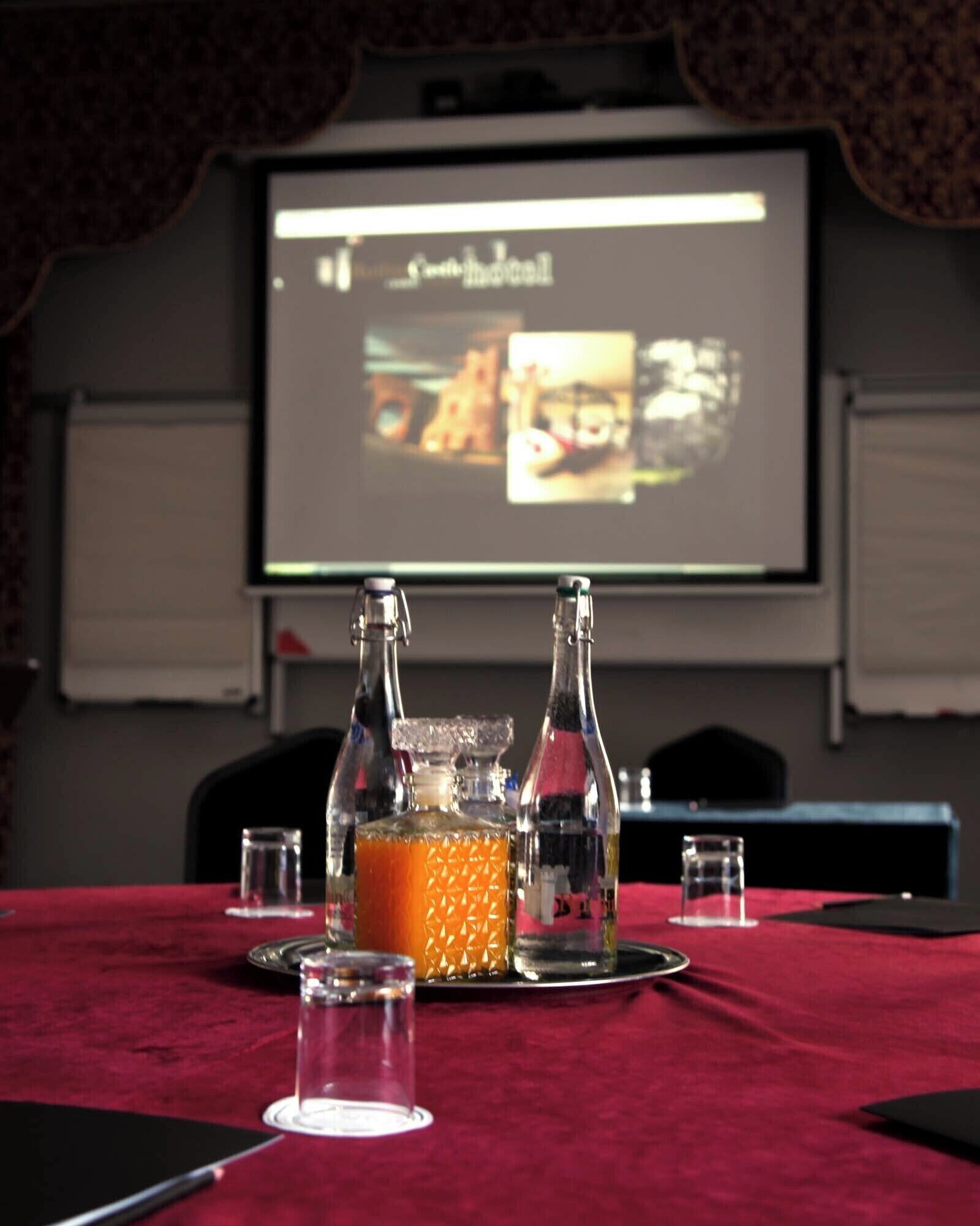 A conference room with a projector screen displaying a presentation, a red tablecloth, two bottles of orange liquid, and several glasses.