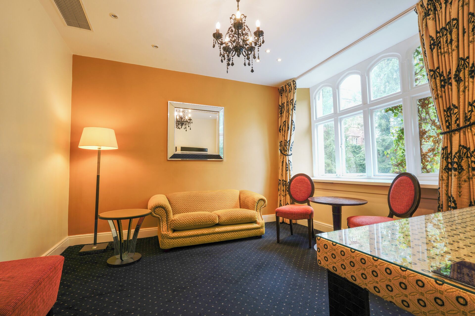 A room with a sofa, two chairs, a small table, a lamp, a chandelier, a mirror, and a window with curtains.