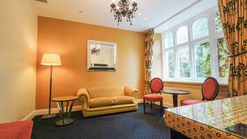 A room with a sofa, two chairs, a small table, a lamp, a chandelier, a mirror, and a window with curtains.