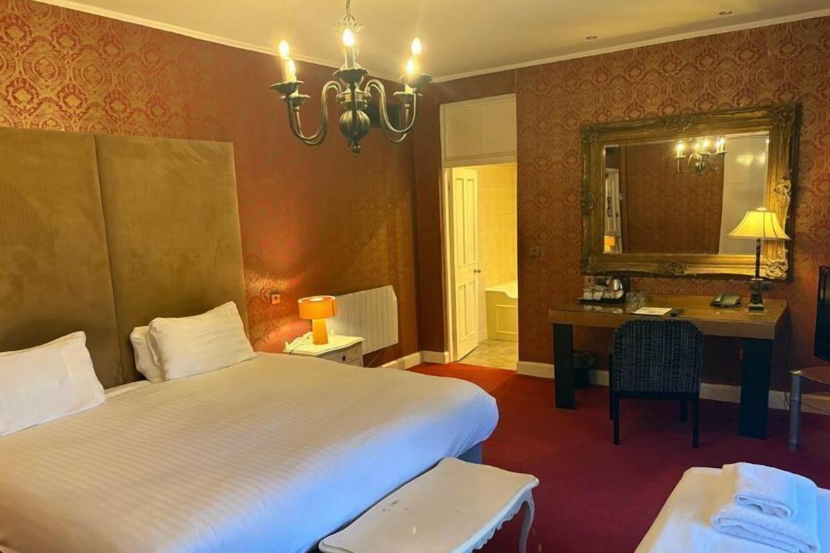 A hotel room with a large bed, a desk with a chair, a mirror, and a chandelier.