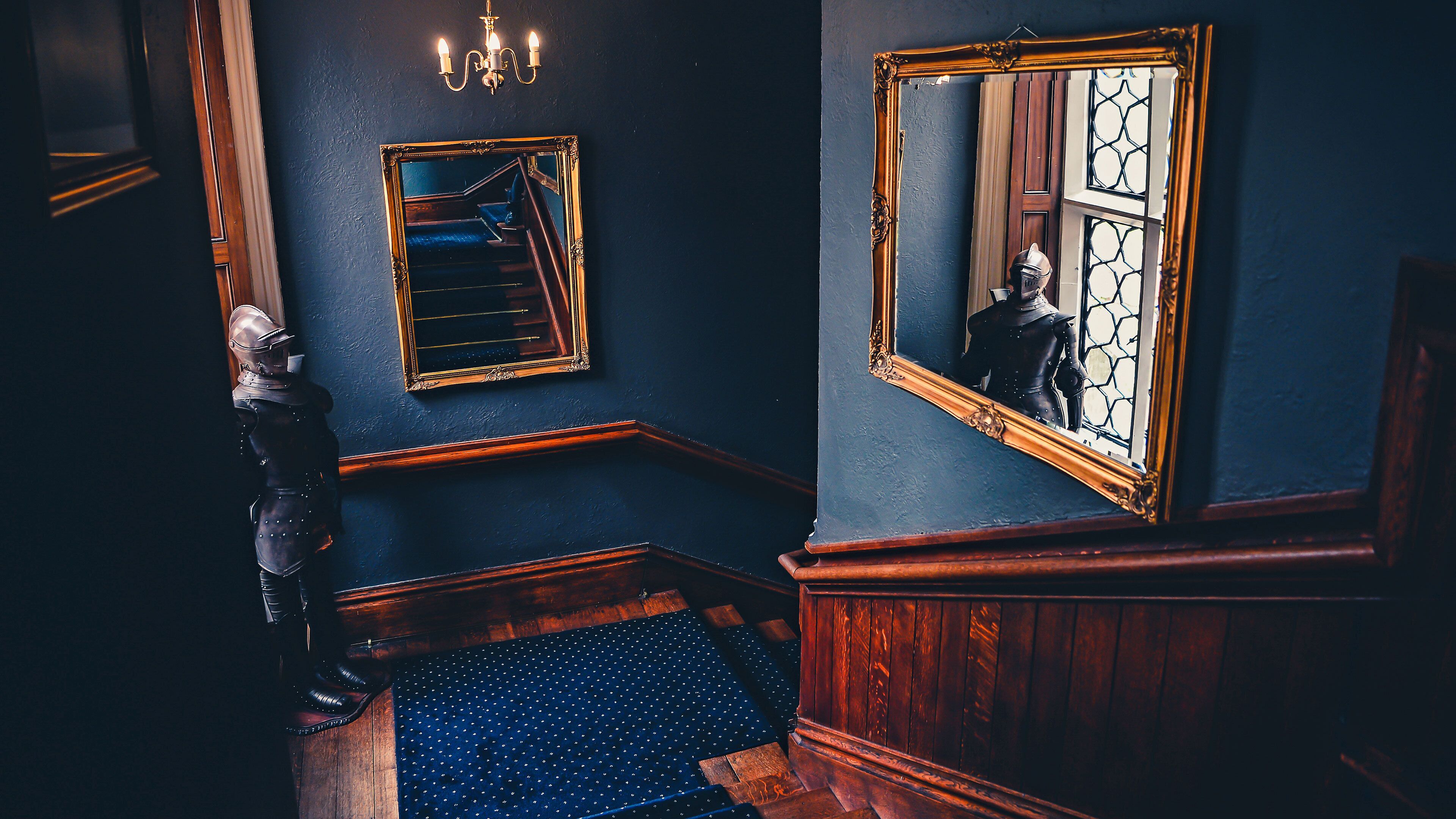 A room with a wooden staircase, a large mirror reflecting a window, and a suit of armor against the wall.