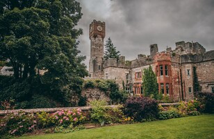 Um castelo histórico com uma torre imponente, vegetação exuberante e um jardim bem cuidado.