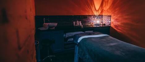 A dimly lit room with a bed, a small table, and a wall-mounted shelf with various items.