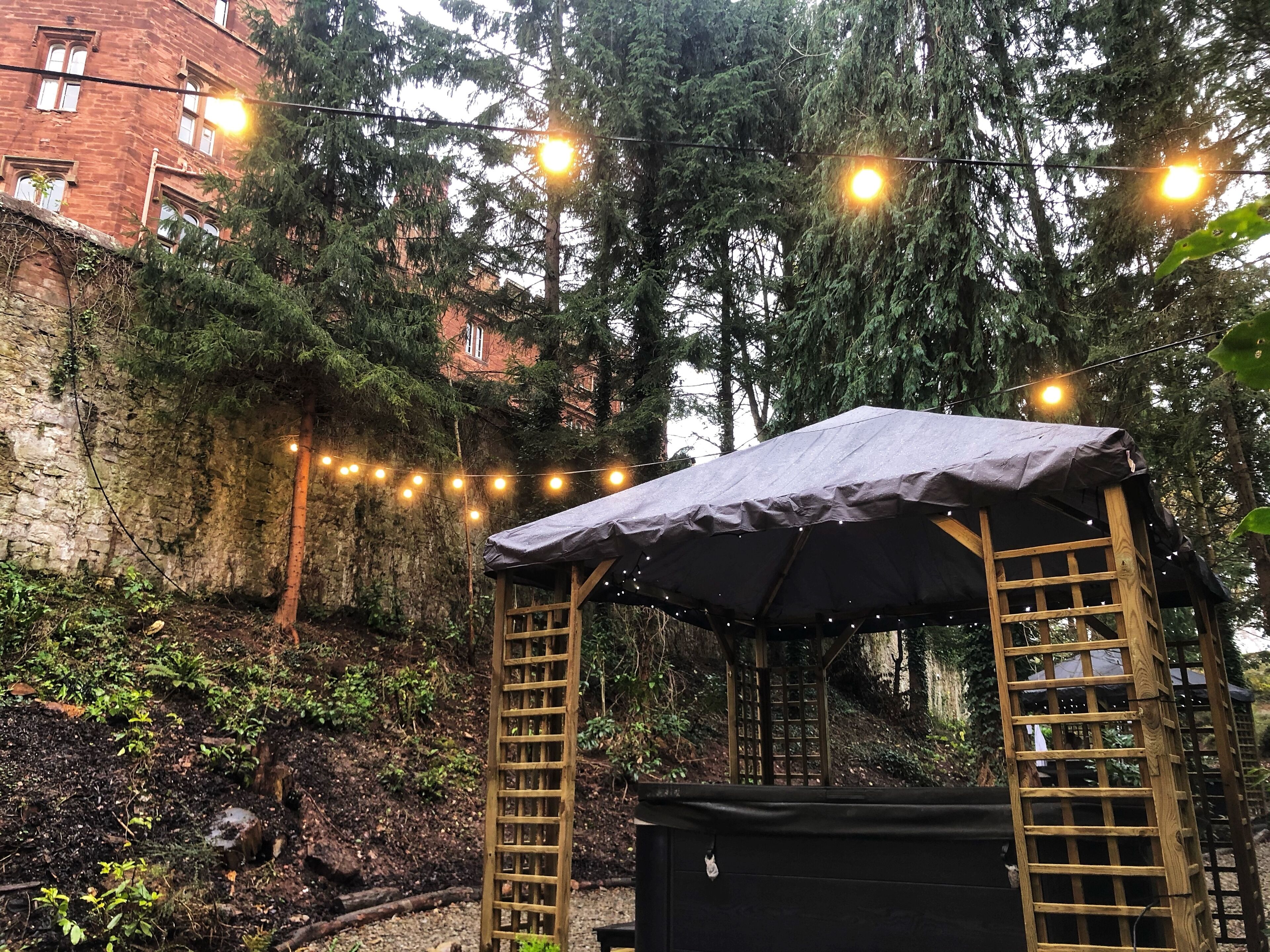 Una pérgola de madera con techo, rodeada de árboles y un muro de piedra.