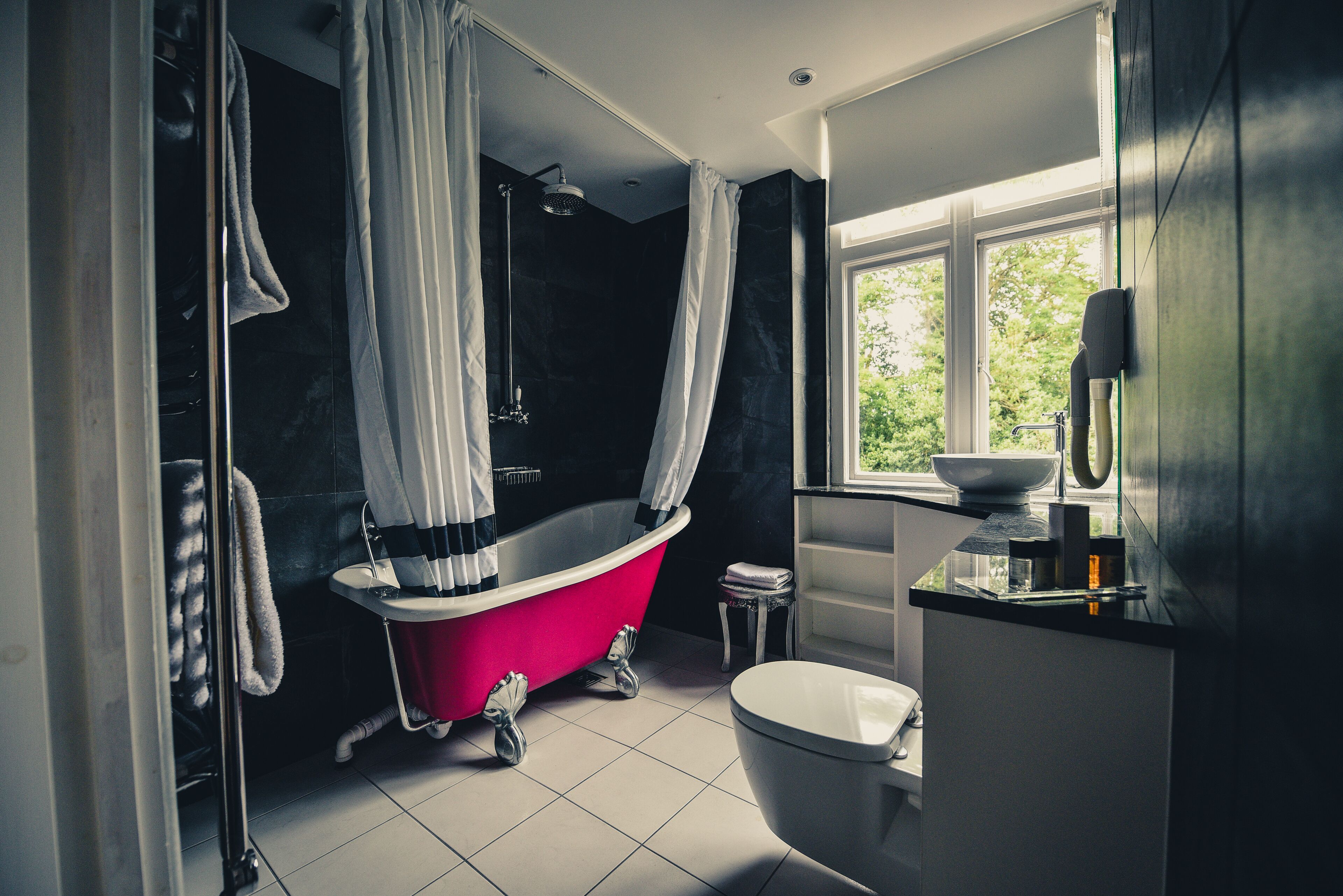 A modern bathroom with a freestanding bathtub, a toilet, and a window providing a view of greenery outside.