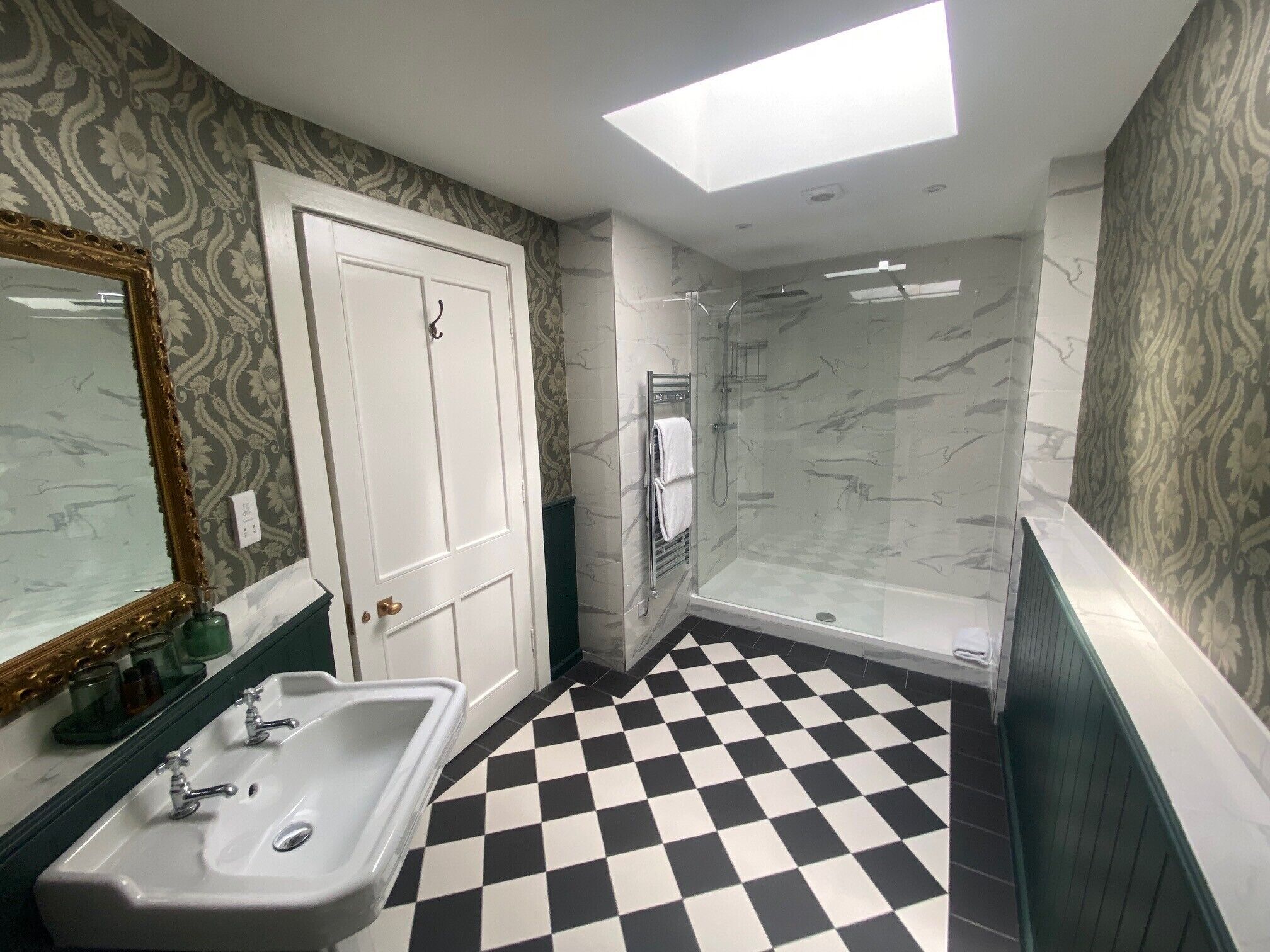 Une salle de bain avec un carrelage  à motifs géométriques, une douche en verre, un lavabo avec deux robinets, un miroir avec un cadre décoratif et un papier peint à motifs.